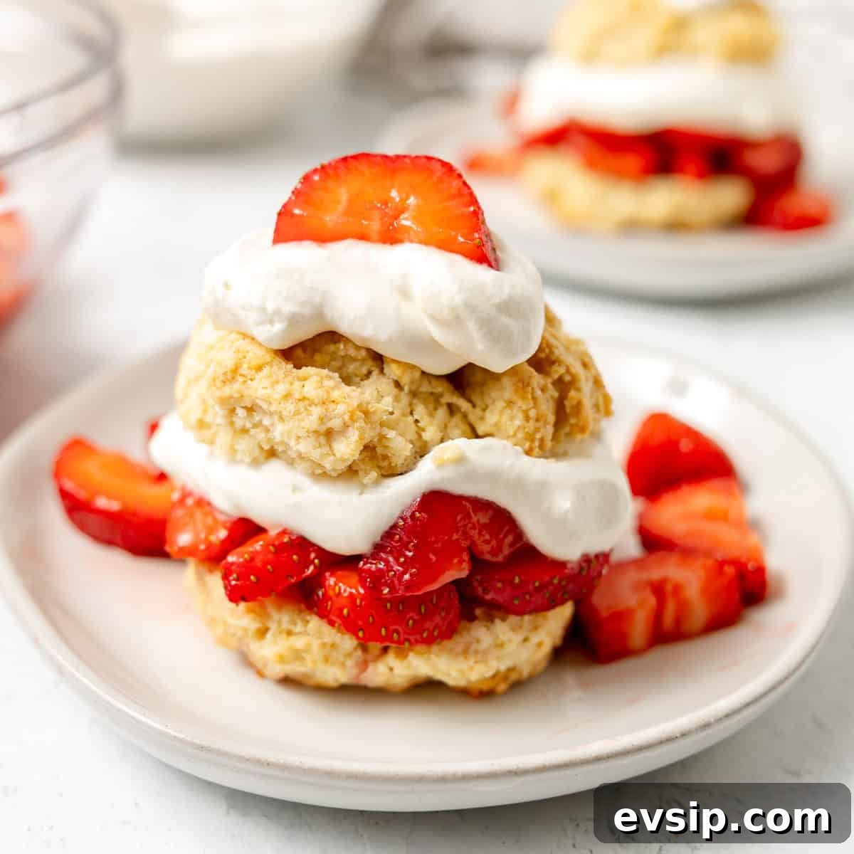 A strawberry shortcake dessert with sliced strawberries and whipped cream layered between two biscuit halves on a white plate.