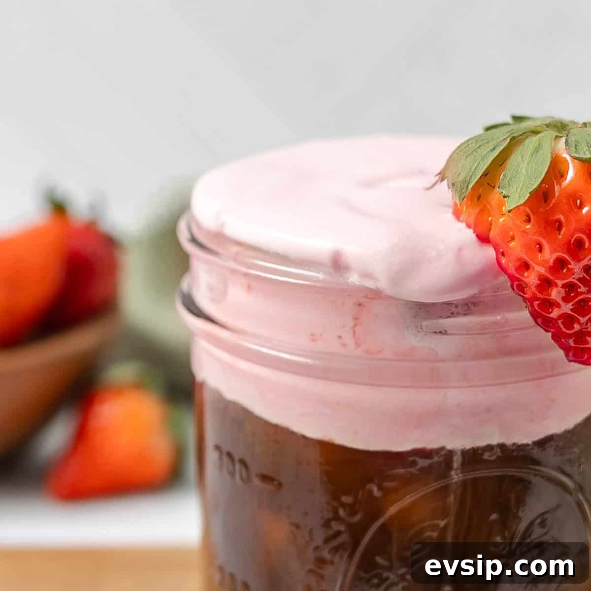 A glass jar filled with iced coffee is topped with a thick layer of strawberry cream and garnished with a fresh strawberry.