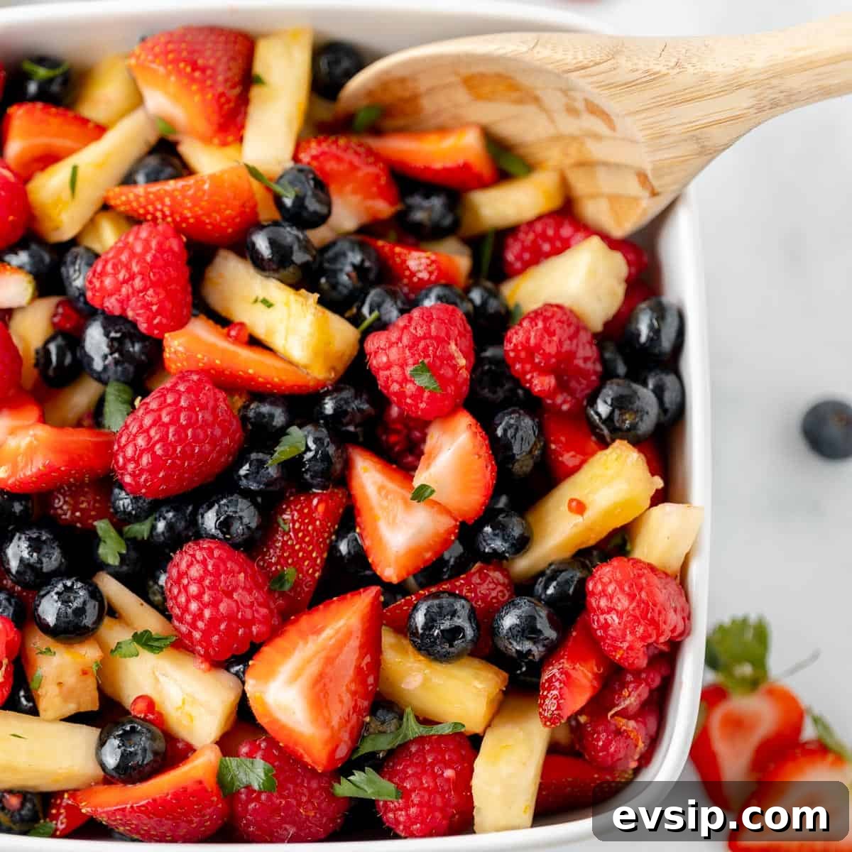 A white bowl filled with a fruit salad containing strawberries, raspberries, blueberries, pineapple chunks, and garnished with herbs. A wooden spoon rests on the side.