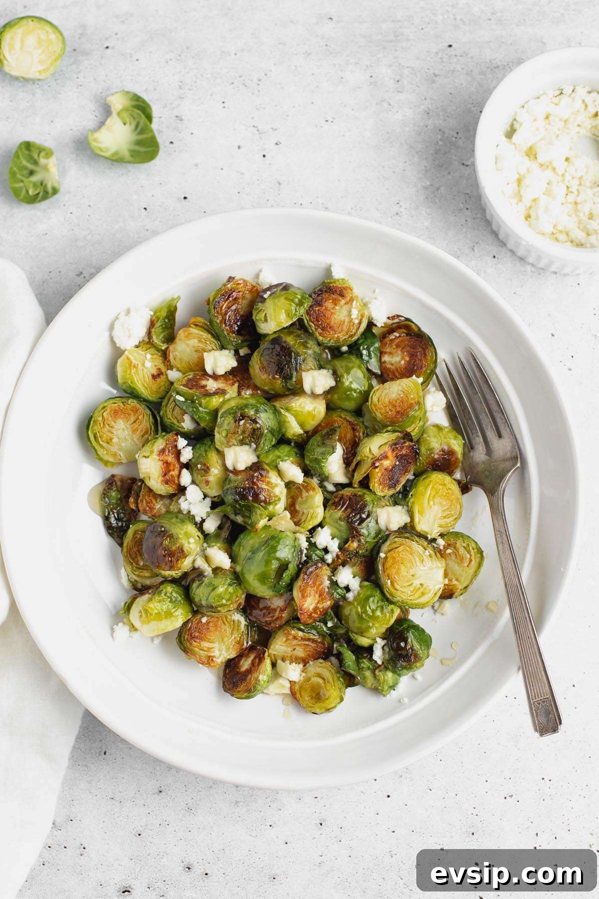 Roasted Brussels Sprouts with Goat Cheese and Honey Glaze 10 Roasted Brussels sprouts with goat cheese on a round plate with a vintage fork with raw Brussels in the background.