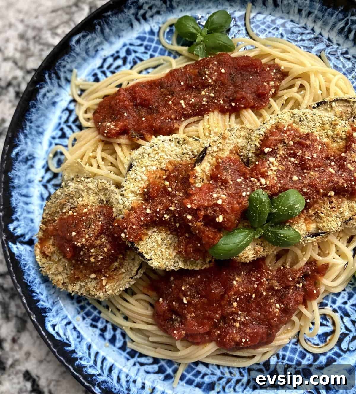 Vegan eggplant parmesan recipe on top of spaghetti with marinara sauce, topped with vegan parmesan and fresh basil on a plate on the table.