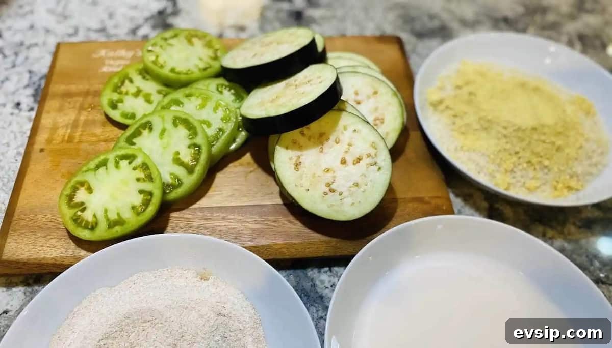 Sliced eggplant and green tomatoes on a cutting board. bowls are around the cutting board for dredging in flour, plant milk, and a nutritional yeast and breadcrumb mixture.
