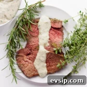 A few slices of herb mustard crusted roast beef on a white plate with creamy herb sauce drizzled over top.