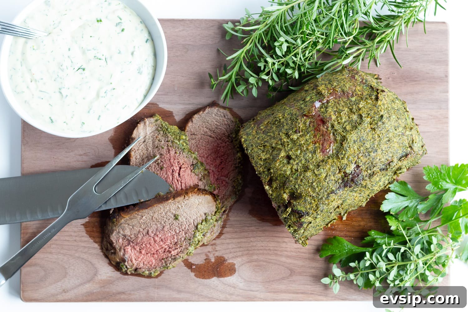 Overhead photo of herb mustard crusted roast beef on a wooden carving board with 3 slices carved off.  There are also fresh herbs, bowl of creamy herb sauce and carving utensils laid out on the board.