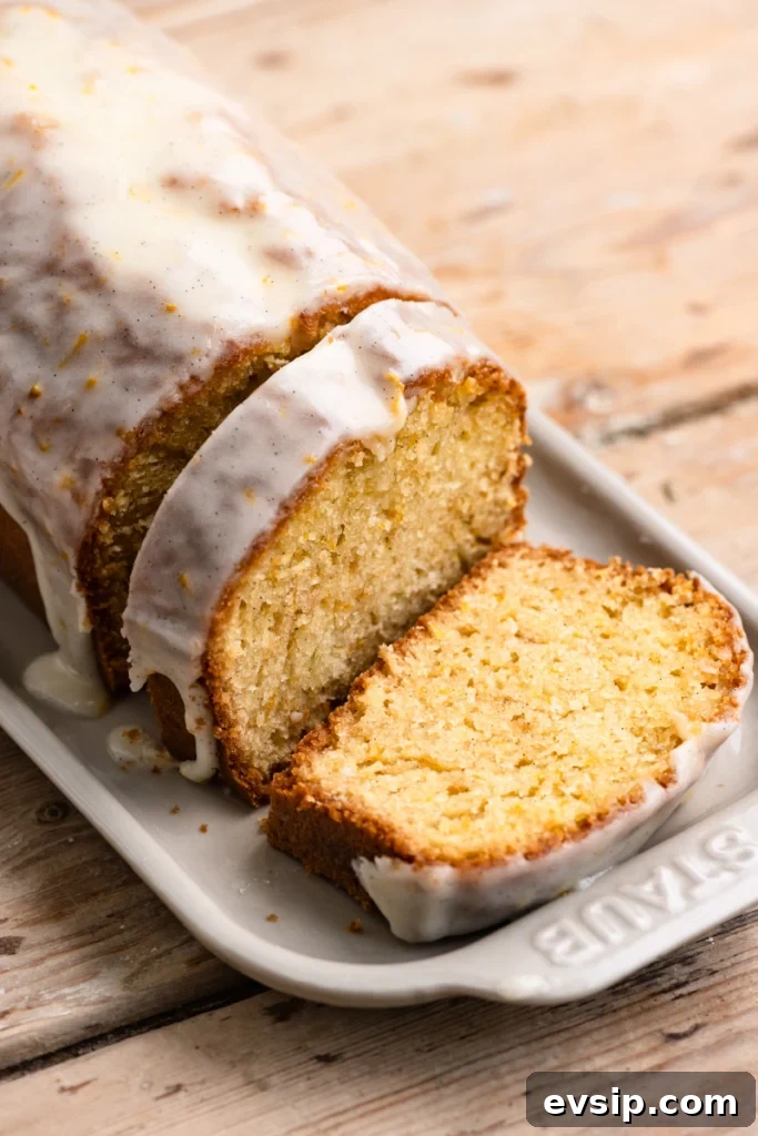 Close up of an orange loaf cake with vanilla glaze sliced on a serving dish with one slice lying flat. 