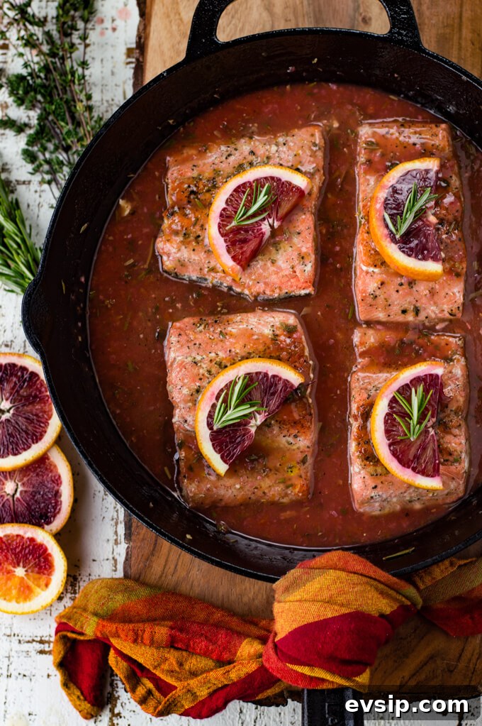 Cast Iron Seared Salmon in Blood Orange Sauce 2 Black cast iron skillet with pieces of salmon sitting in a blood orange sauce. Each salmon has a half slice of blood orange and a small sprig of rosemary on top. Slices of blood oranges and herbs sit next to the pan.