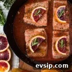 Black cast iron skillet with pieces of salmon sitting in a blood orange sauce. Each salmon has a half slice of blood orange and a small sprig of rosemary on top. Slices of blood oranges and herbs sit next to the pan.