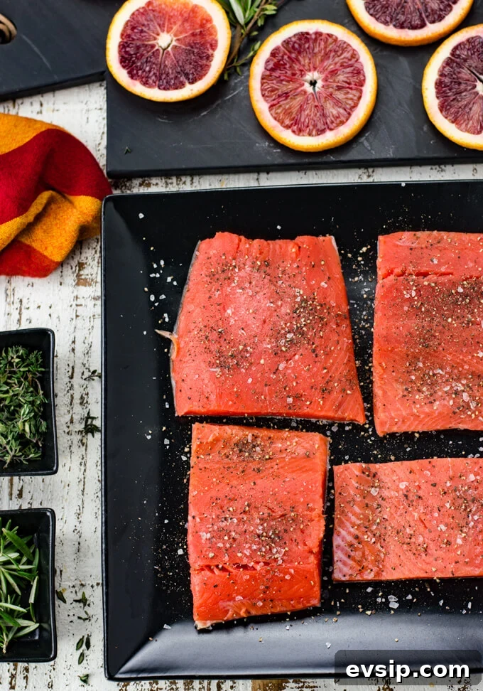 Cast Iron Seared Salmon in Blood Orange Sauce 4 Four pieces of salmon sprinkled with salt and pepper sit on a black plate. Slices of blood oranges sit on a black cutting board above the salmon in the photo. A red and orange napkin and 2 small black bowls with thyme and rosemary sit next to the salmon.