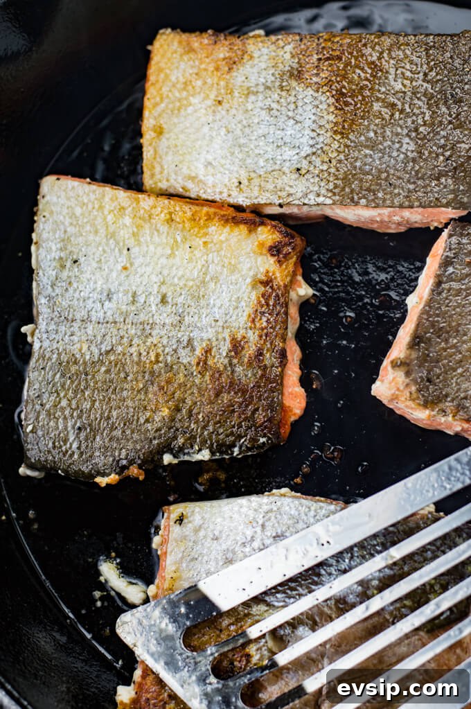 Cast Iron Seared Salmon in Blood Orange Sauce 6 Four pieces of salmon are cooking in a black cast iron skillet with the skins up. A Fish spatula sits on top of them.