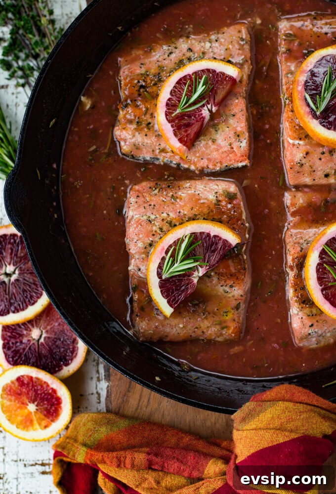 Cast Iron Seared Salmon in Blood Orange Sauce 10 Black cast iron skillet with pieces of salmon sitting in a blood orange sauce. Each salmon has a half slice of blood orange and a small sprig of rosemary on top. Slices of blood oranges and herbs sit next to the pan.