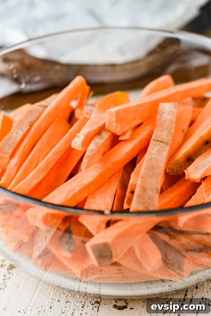 A glass bowl of raw sweet potato fries and two stacked cooking sheets with parchment paper sit in the background.