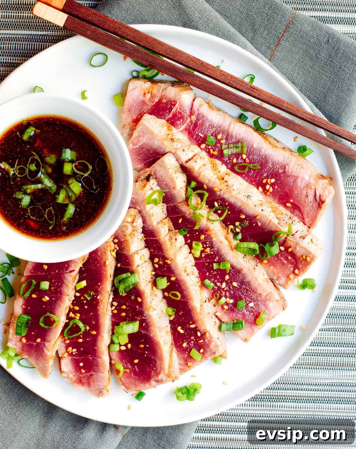 A plate of seared tuna slices garnished with chopped green onions and sesame seeds, accompanied by a small bowl of soy-based dipping sauce. Chopsticks rest on the edge of the plate. The plate is set on a blue-gray placemat.