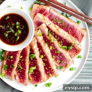 A plate of grilled tuna slices garnished with chopped green onions and a sprinkle of seasoning. There is a small bowl of dipping sauce with sliced green onions on the side. Chopsticks rest on the edge of the plate. The background includes a textured cloth and mat.