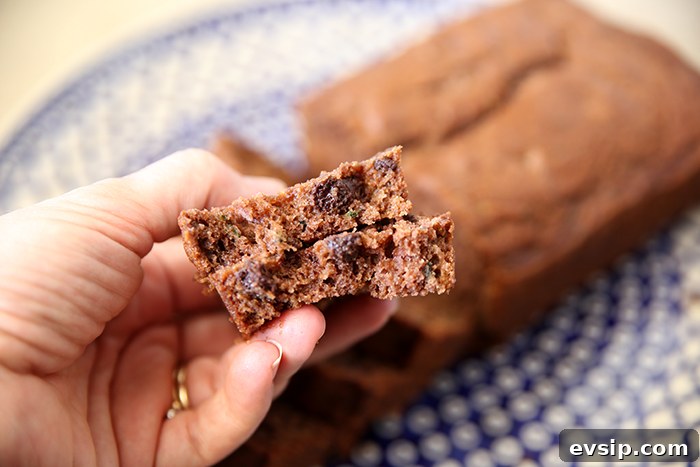 A slice of chocolate zucchini bread broken in half held above a dark blue Polish pottery plate with a bubble pattern in the middle and a lattice pattern around the edge with a loaf of chocolate zucchini bread partially sliced.