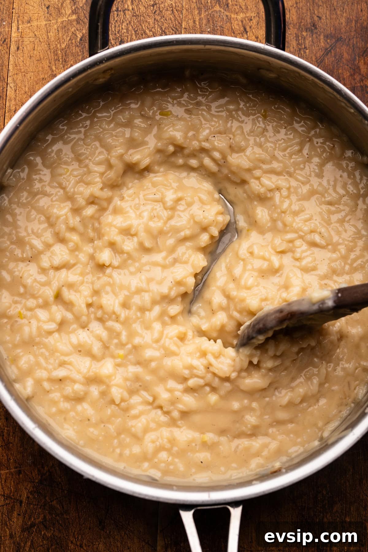 A wooden spoon stirring creamy risotto in a stainless steel pan on a wooden background.