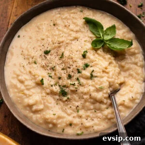 A dark gray bowl with creamy risotto in it with fresh basil on top of it and a fork in the bowl. The bowl of risotto is arranged on a wooden board with white wine, a block of Parmigiano Reggiano cheese, and a gray napkin placed around it.