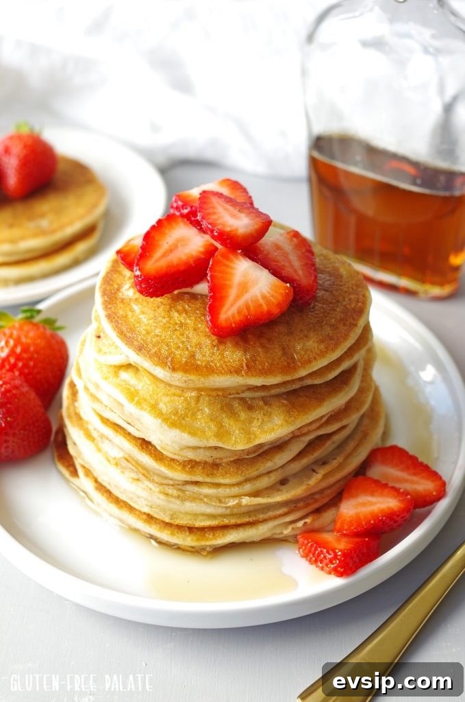 Irresistible Gluten-Free Brunch Recipes You’ll Crave Again 6 Gluten-Free Pancakes stacked on a white plate, topped with strawberries next to a jar of maple syrup.