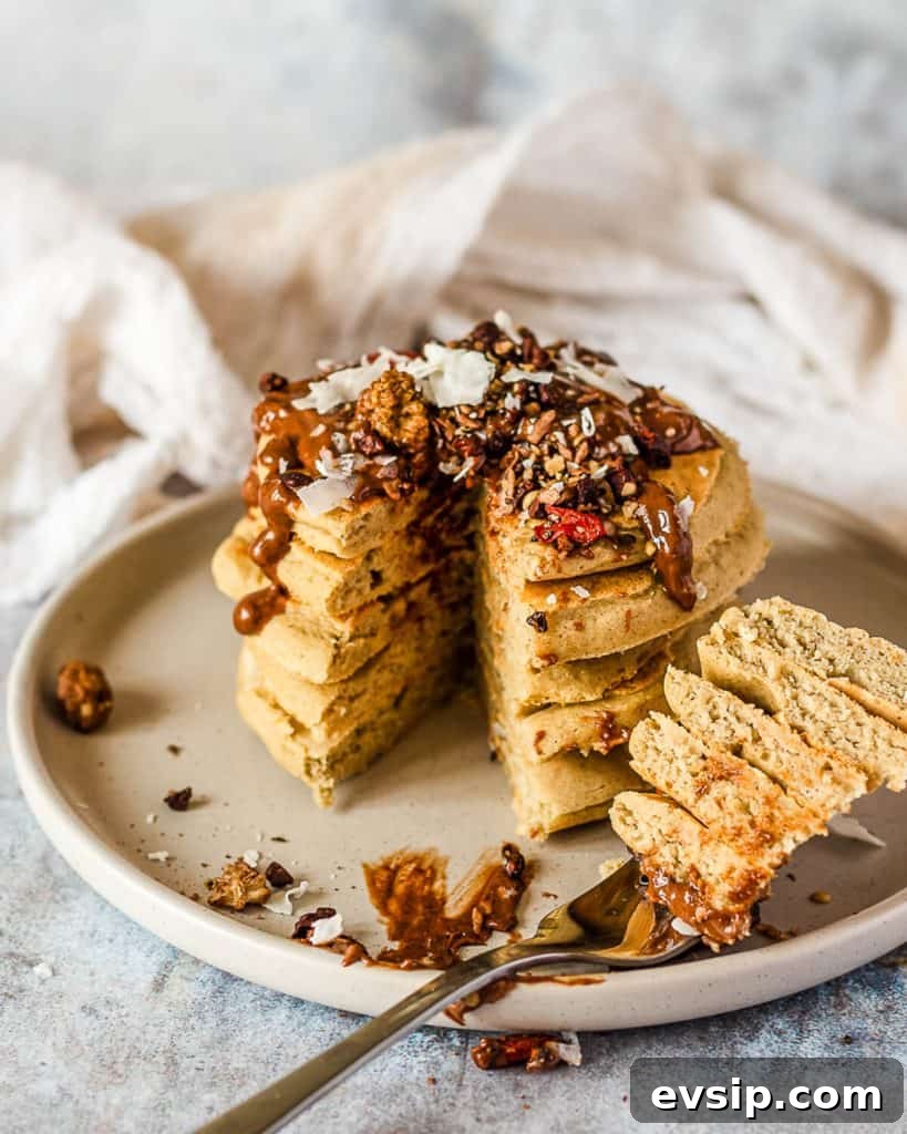 A fork holding a bite of vegan buckwheat pancake in front of a stack of pancakes topped with cacao hazelnut butter and seeds.