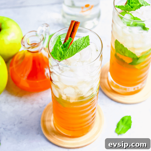 Two tall glasses of iced orange drink with mint leaves and cinnamon sticks, next to green apples.