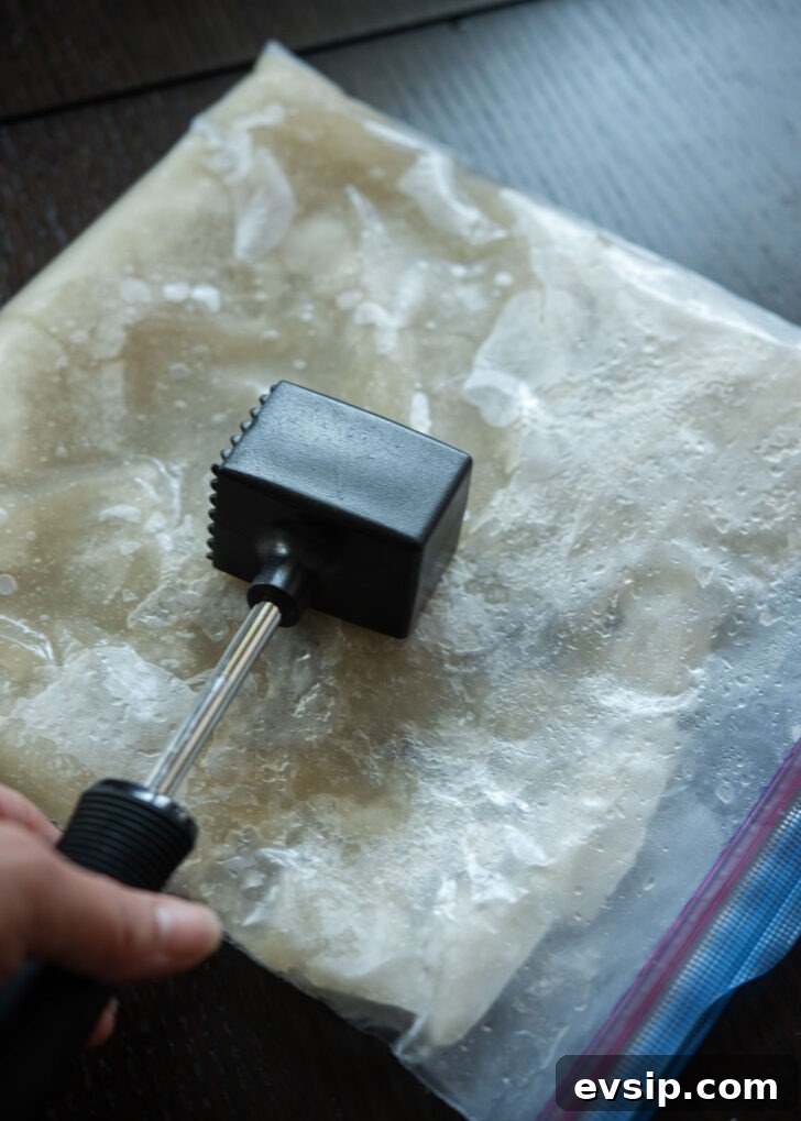 Partially frozen naengmyeon broth slab being crushed with a meat hammer to create a slushy texture.