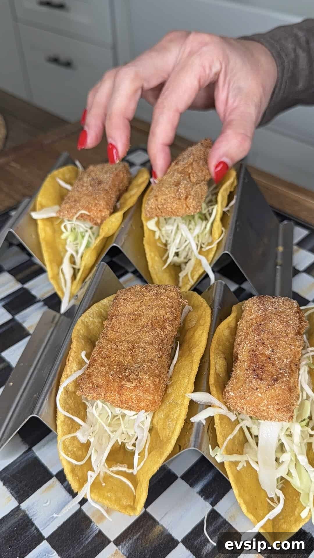 Crispy Mahi Mahi Fish Tacos with Zesty Slaw 11 A hand with red nail polish places a piece of breaded fish on a taco filled with shredded cabbage. Three other similar fish tacos sit in metal taco holders on a checkered surface.