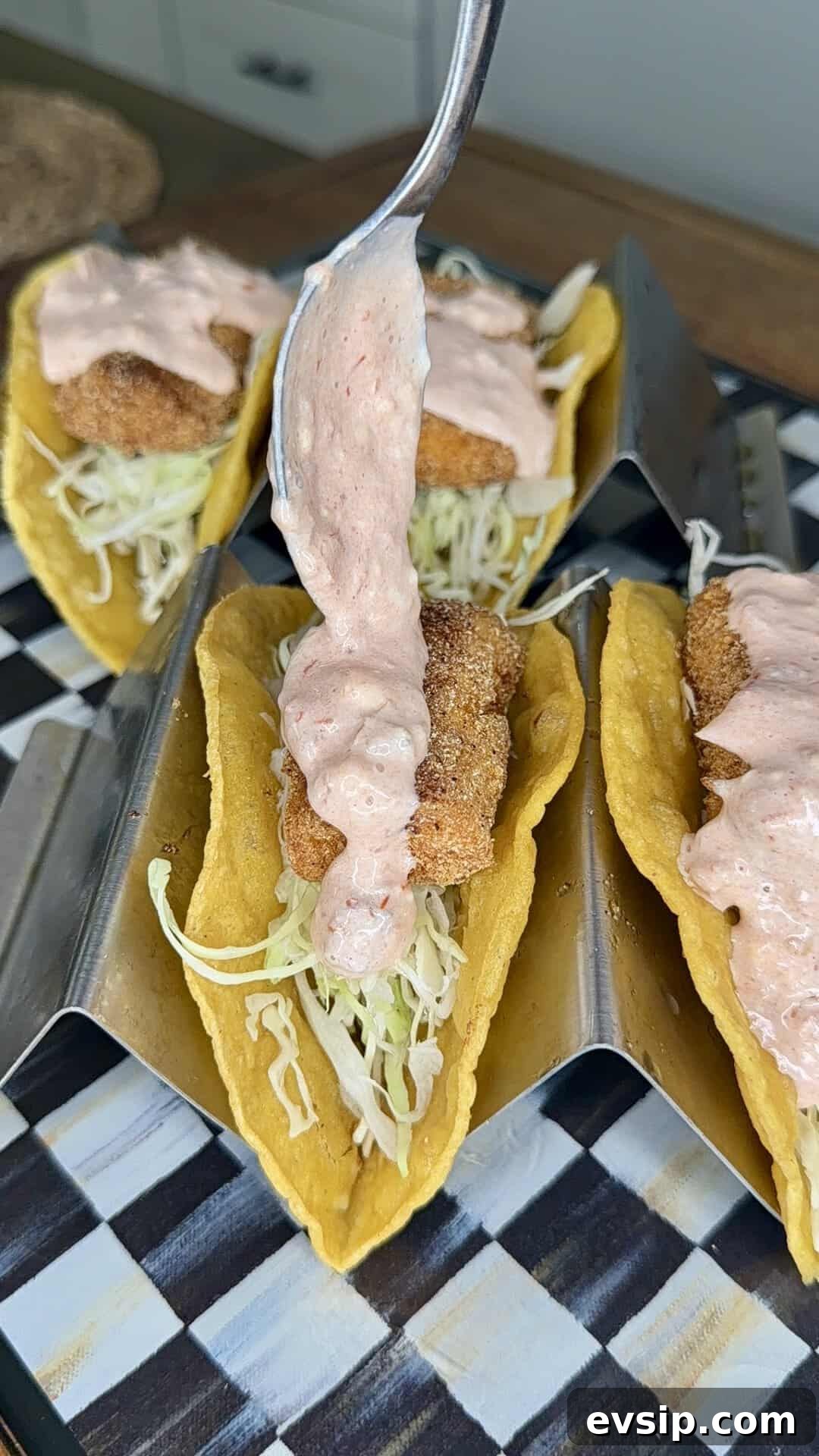 Crispy Mahi Mahi Fish Tacos with Zesty Slaw 15 A spoon drizzles creamy sauce over crispy fish tacos filled with shredded lettuce in yellow corn tortillas, arranged in a metal taco holder on a checkered tray.