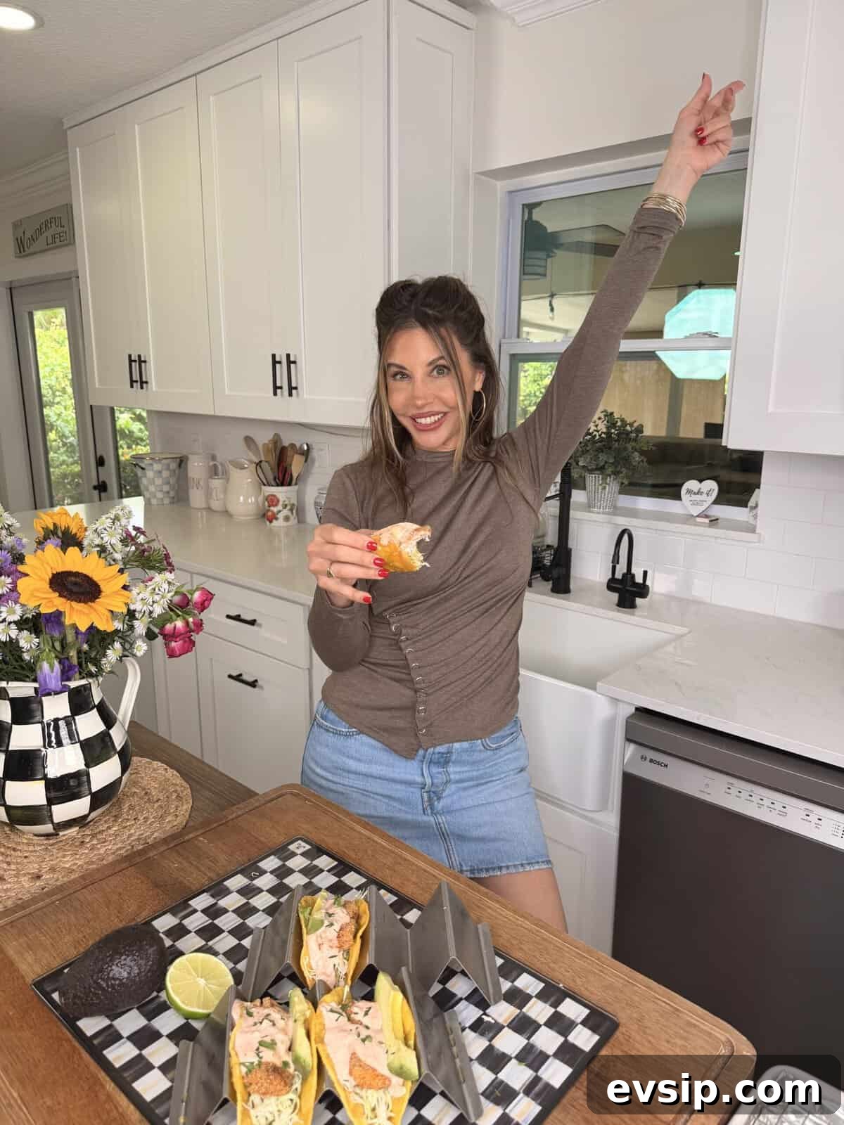 Crispy Mahi Mahi Fish Tacos with Zesty Slaw 18 A woman in a brown top and denim skirt stands in a bright kitchen, smiling and pointing upward with one hand while holding a taco in the other. On the counter are tacos, a half avocado, a lime, and a vase of flowers.