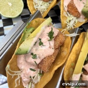Crispy Mahi Mahi Fish Tacos with Zesty Slaw 20 Three crispy fish tacos in taco holders, topped with shredded cabbage, sliced avocado, creamy pink sauce, and chopped herbs. A halved lime and part of an avocado are visible in the background.