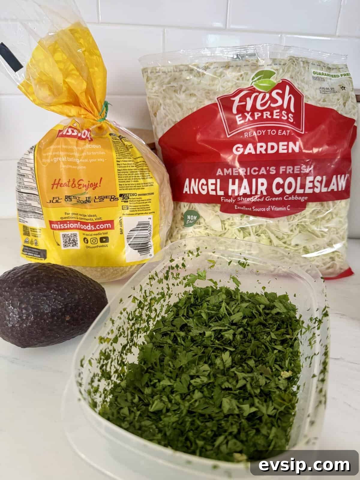 Crispy Mahi Mahi Fish Tacos with Zesty Slaw 3 A bag of tortillas, a bag of angel hair coleslaw, a whole avocado, and a container of chopped cilantro are arranged on a white kitchen counter.