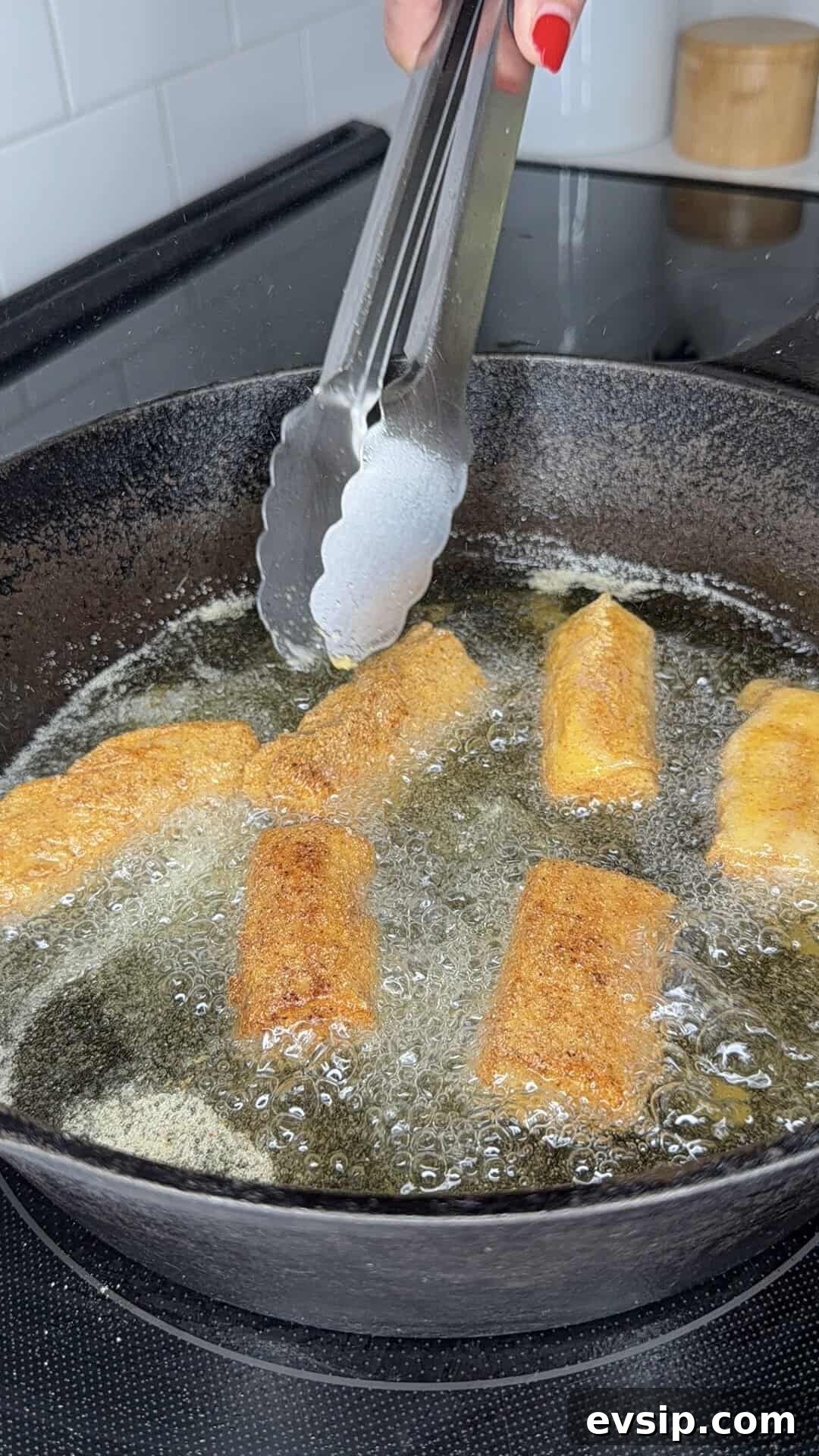 Crispy Mahi Mahi Fish Tacos with Zesty Slaw 6 A hand using metal tongs to fry golden, breaded rolls in hot oil inside a black cast iron skillet on a stovetop.