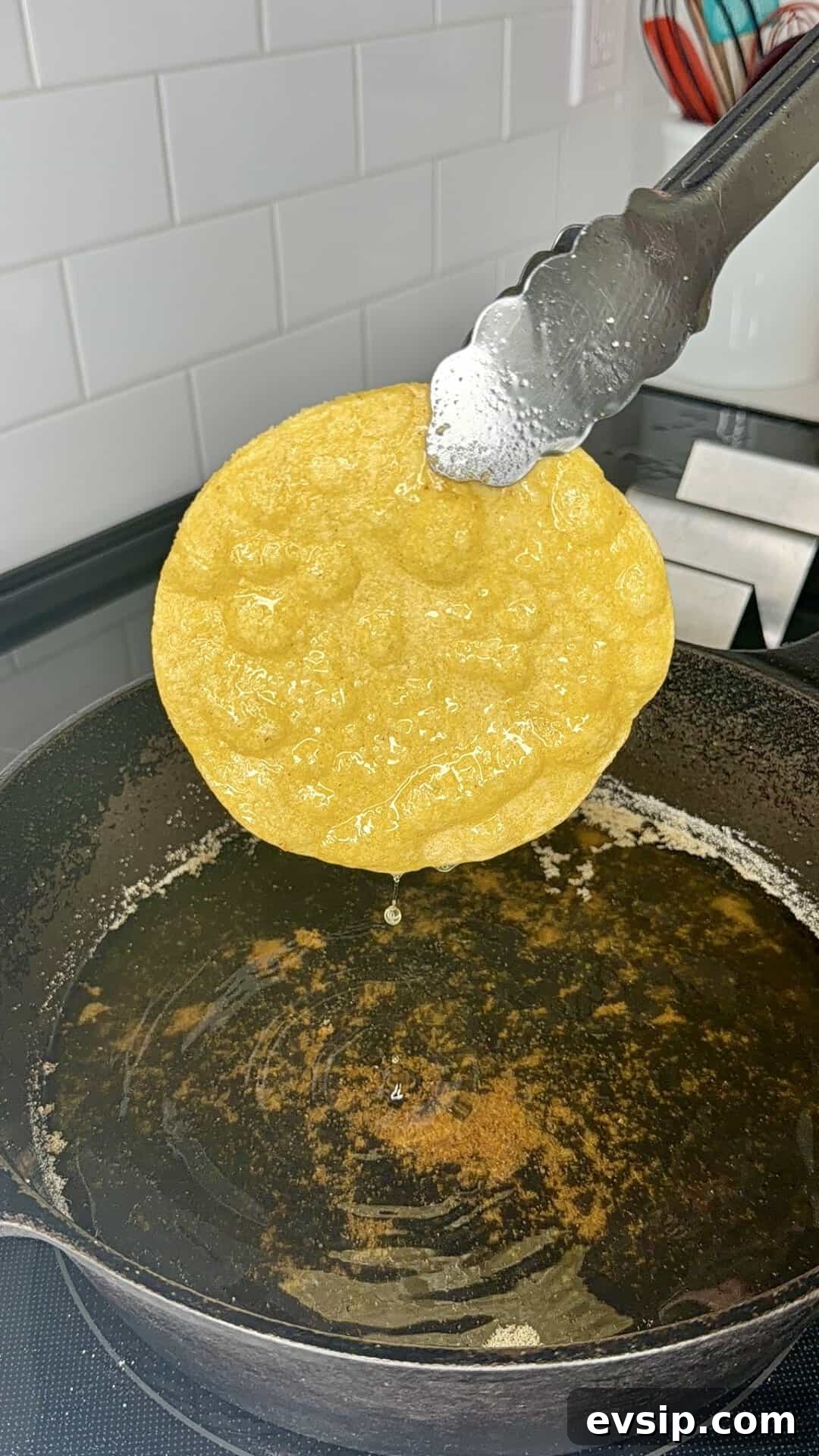 Crispy Mahi Mahi Fish Tacos with Zesty Slaw 8 A hand holds metal tongs lifting a yellow corn tortilla from hot oil in a black cast iron skillet on a stovetop, with droplets of oil dripping from the tortilla.