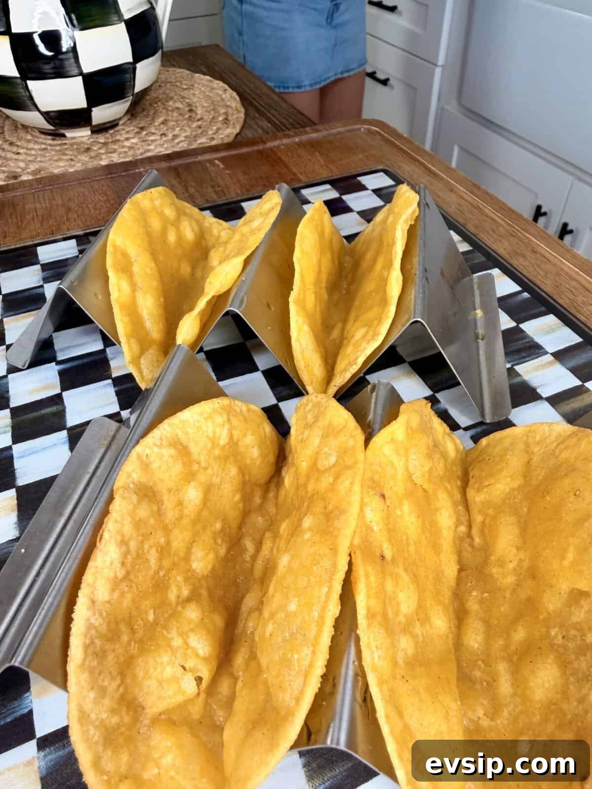 Crispy Mahi Mahi Fish Tacos with Zesty Slaw 9 Four crunchy taco shells sit in metal taco holders on a black-and-white checkered surface, with part of a person and a kitchen background visible.