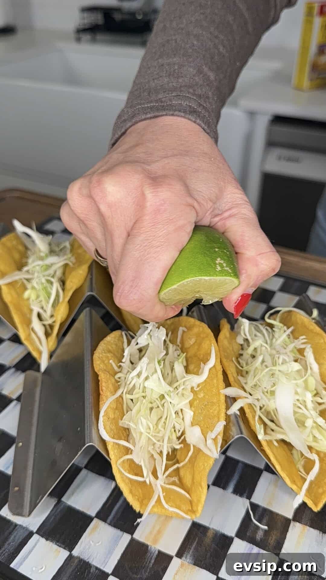 Crispy Mahi Mahi Fish Tacos with Zesty Slaw 10 A hand squeezes a lime wedge over hard-shell tacos filled with shredded cabbage, arranged on a taco holder on a checkered surface.