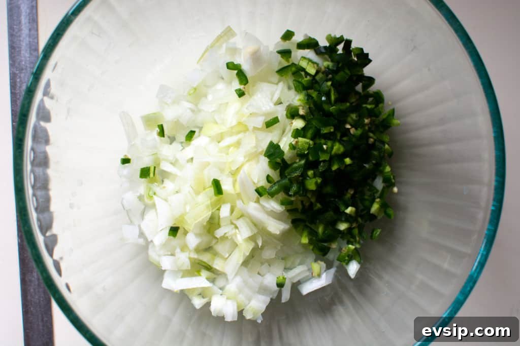 A glass bowl with chopped onion and chopped jalapeño pepper on a white counter. 