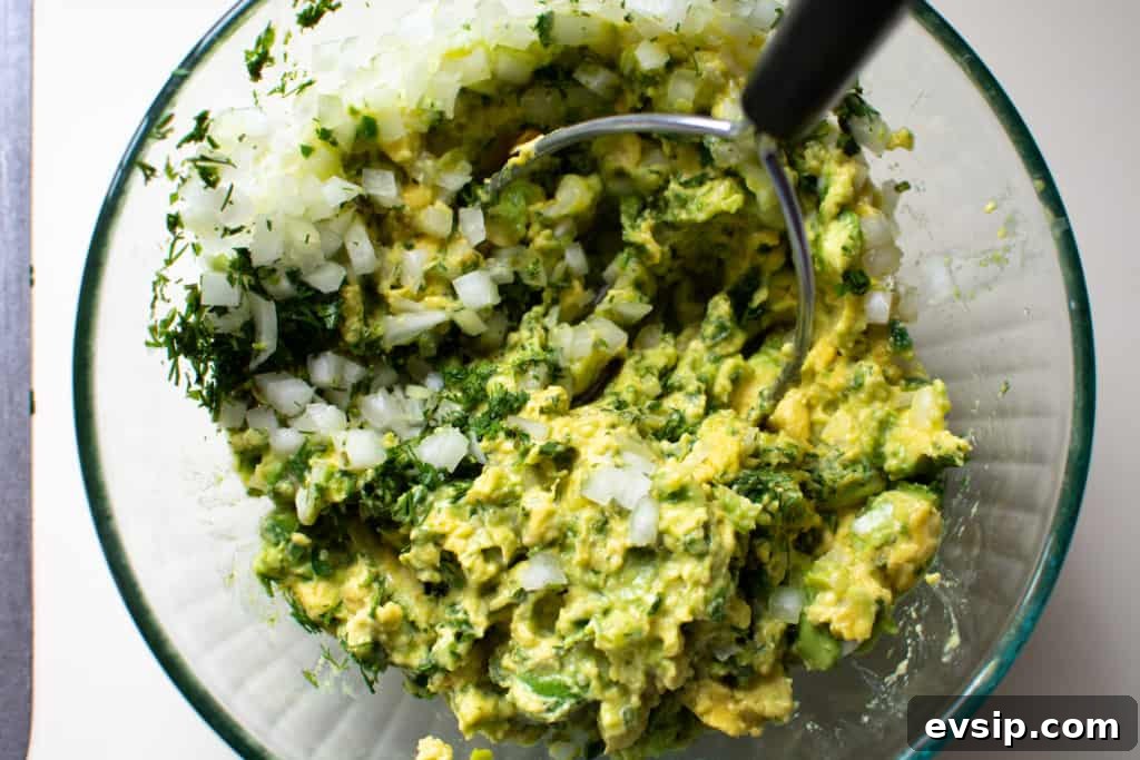 A glass bowl filled with mashed avocado, chopped white onion, and minced cilantro with a potato masher in it. 