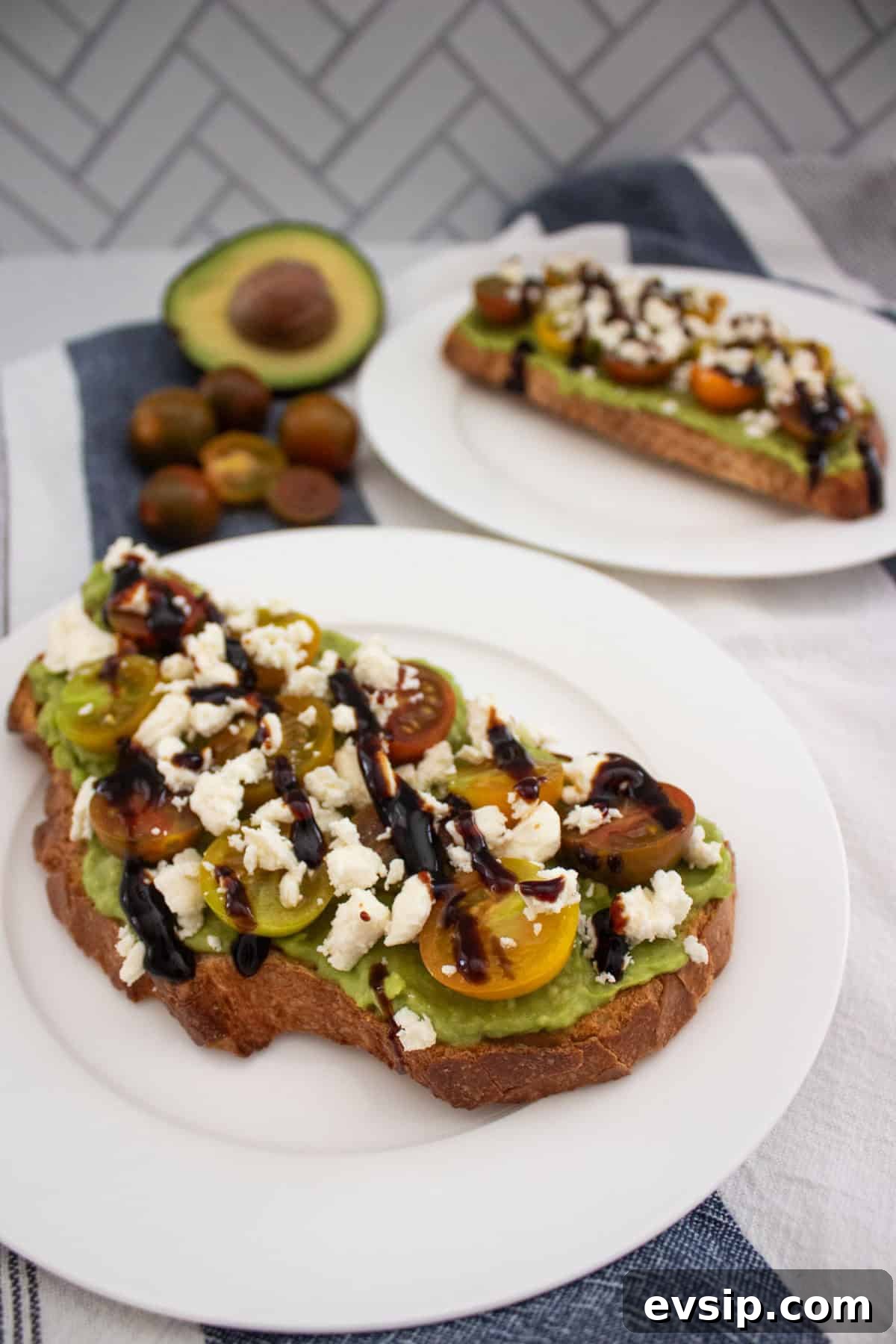 A slice of toast topped with mashed avocado, halved cherry tomatoes, crumbled cheese, and a drizzle of balsamic glaze on a white plate. Another similar toast, avocado, and tomatoes are in the background.