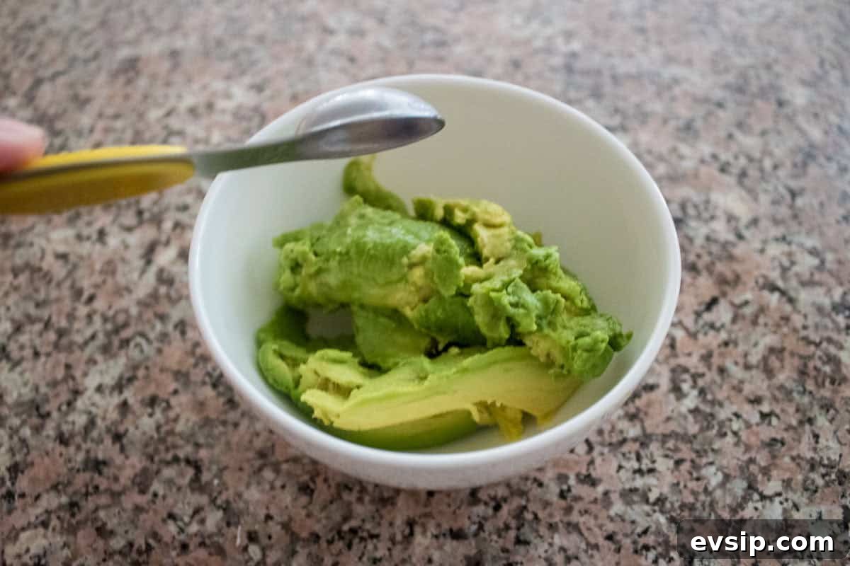 A bowl containing mashed avocado sits on a granite countertop, with a spoon resting on the edge of the bowl.