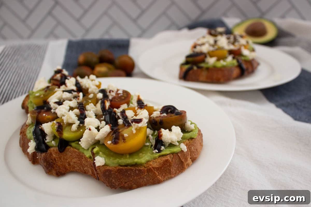 Sliced bread topped with smashed avocado, halved tomatoes, feta cheese, and balsamic glaze on a white plate, with more toast and an avocado in the background.