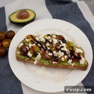 A slice of toast topped with mashed avocado, halved cherry tomatoes, crumbled feta, and a drizzle of balsamic glaze on a white plate. An avocado half and some cherry tomatoes sit nearby on a striped cloth.