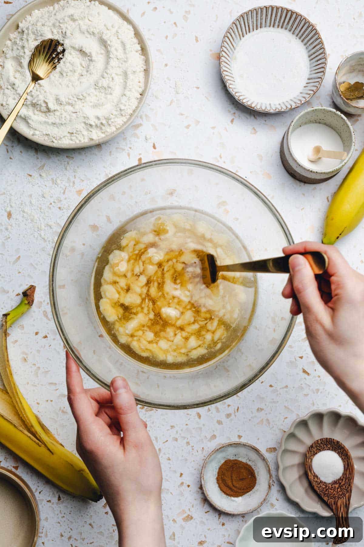 hand using a fork to mix oil, mashed banana, and honey in a bowl. 