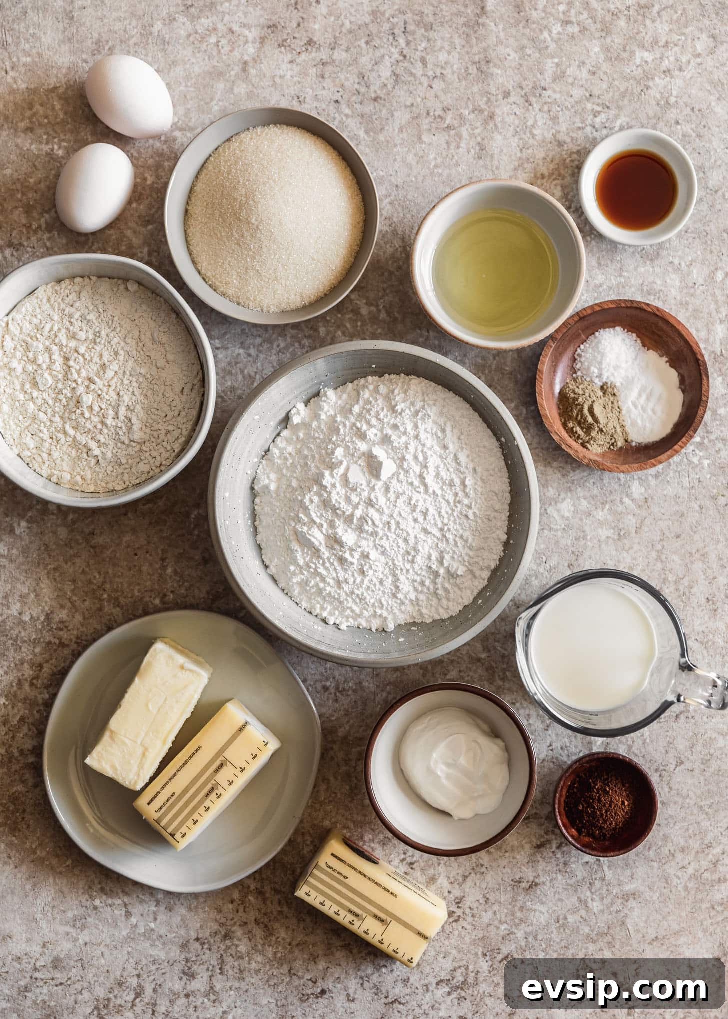 White and wood bowls of baking ingredients like flour, sugar, butter, spices, milk, vanilla, and sour cream on a beige counter.