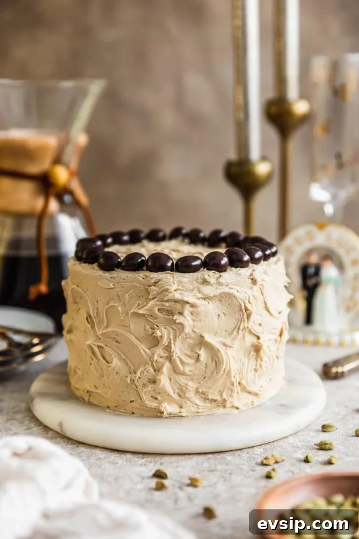 Cardamom cake with coffee buttercream on a marble plate next to a wood bowl of cardamom pods, white linen, Chemex, and wedding cake topper on a beige background.