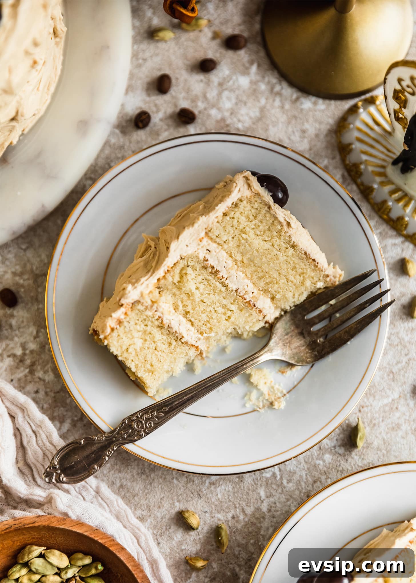 A white plate of cardamom cake with coffee frosting on a beige counter next to a white linen, wedding cake topper, coffee beans, and cardamom pods.