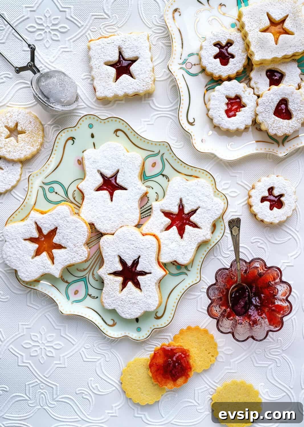 Assorted Austrian Linzer cookies with powdered sugar and jam filling are arranged on decorative plates, alongside a jar of jam and a sifter of powdered sugar on a white embroidered tablecloth.