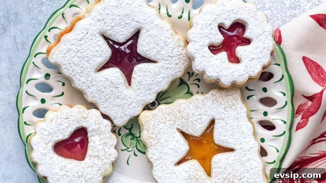 A plate of classic linzer cookies with powdered sugar on top, each featuring a unique cut-out shape that reveals colorful jam. More linzer cookies and a red-patterned napkin sit nearby on the table.