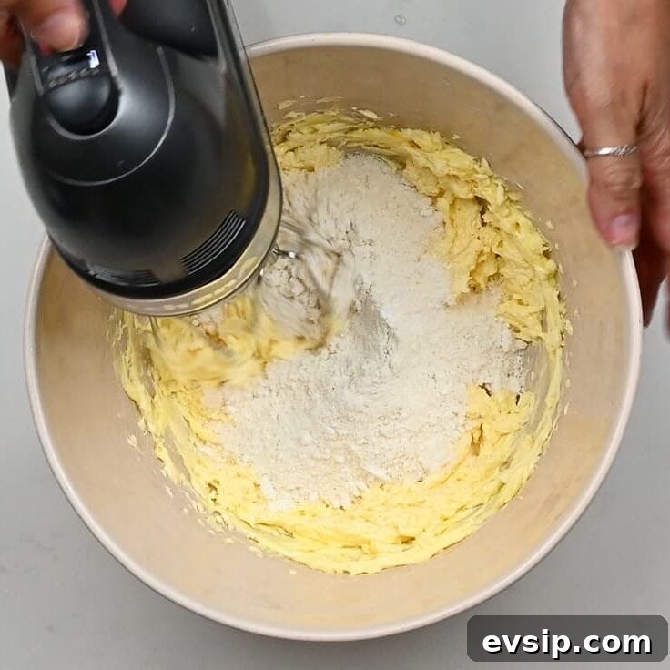 A person uses a hand mixer to blend flour into a yellow batter for linzer cookies in a large mixing bowl, seen from above.