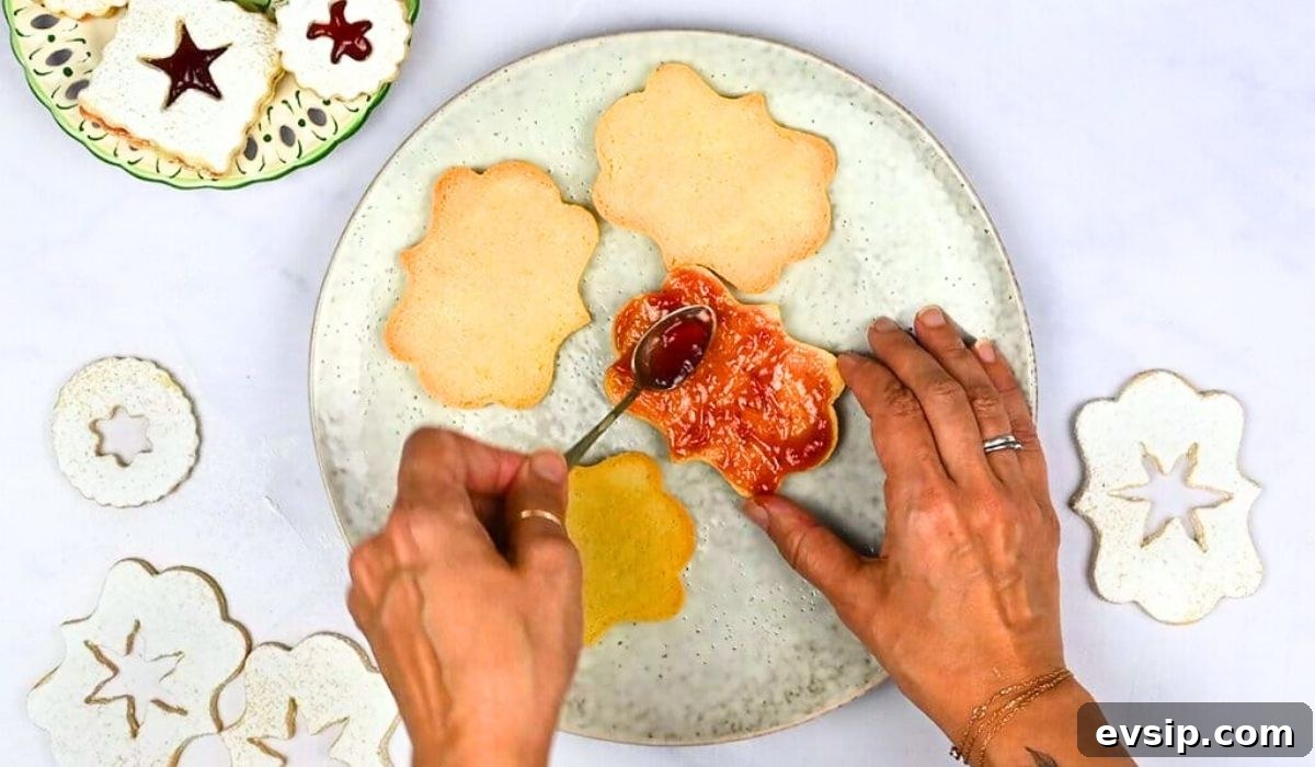 A person spreads red jam onto a flower-shaped cookie with a spoon, preparing Linzer Cookies on a white speckled plate. Decorated cookies and cutouts from this classic linzer cookie recipe are visible nearby on a white surface.