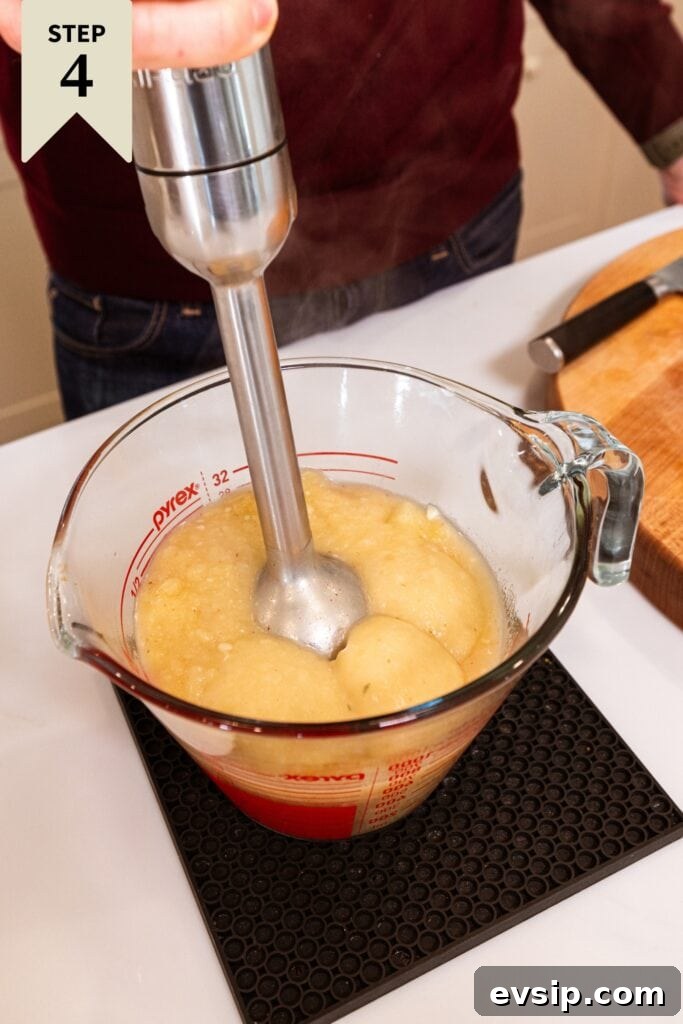 Hand using immersion blender to purée pieces of potato in potato soup to be creamy.