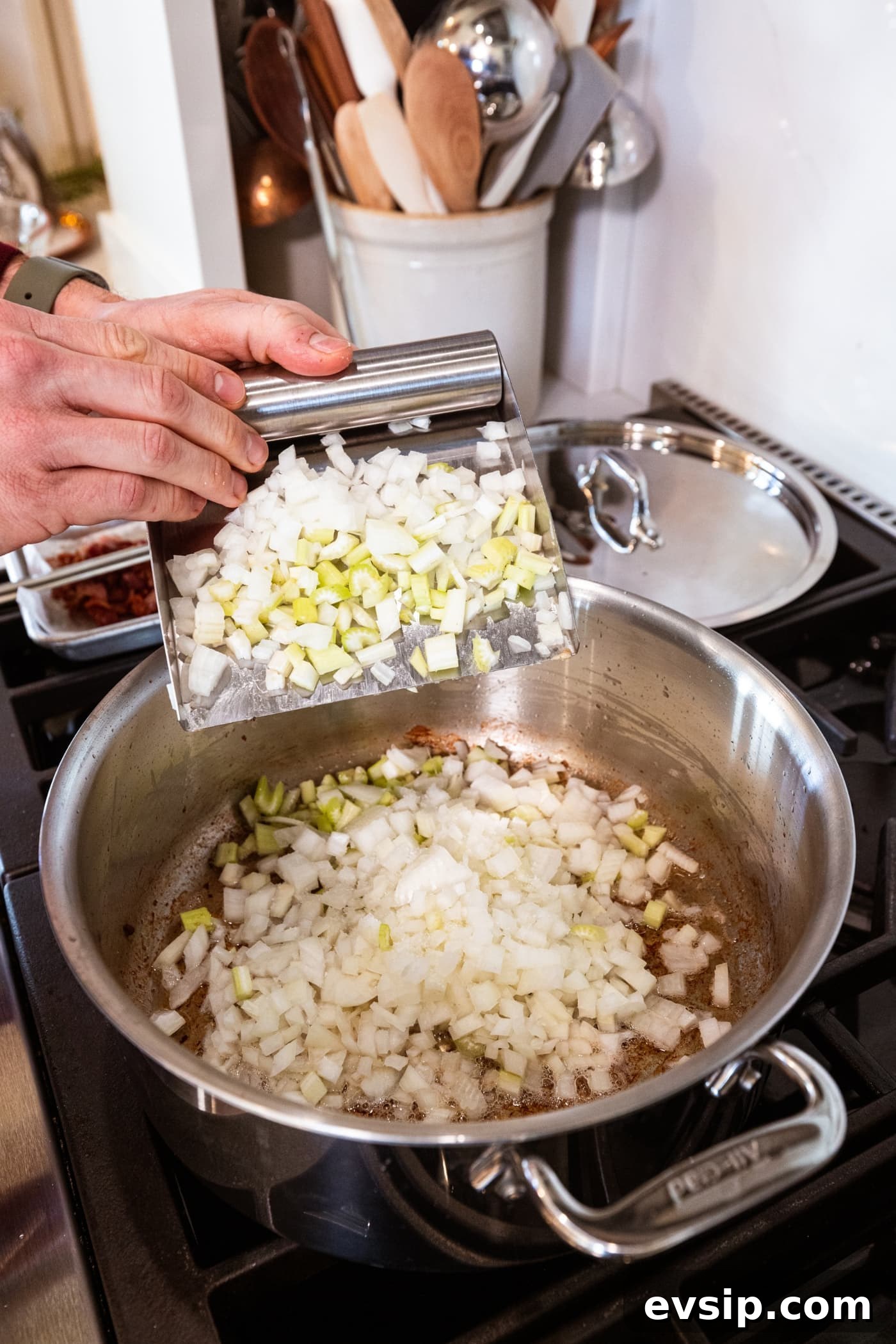 Hand using a scraper to throw diced onion and celery into a large stockpot on the stove.