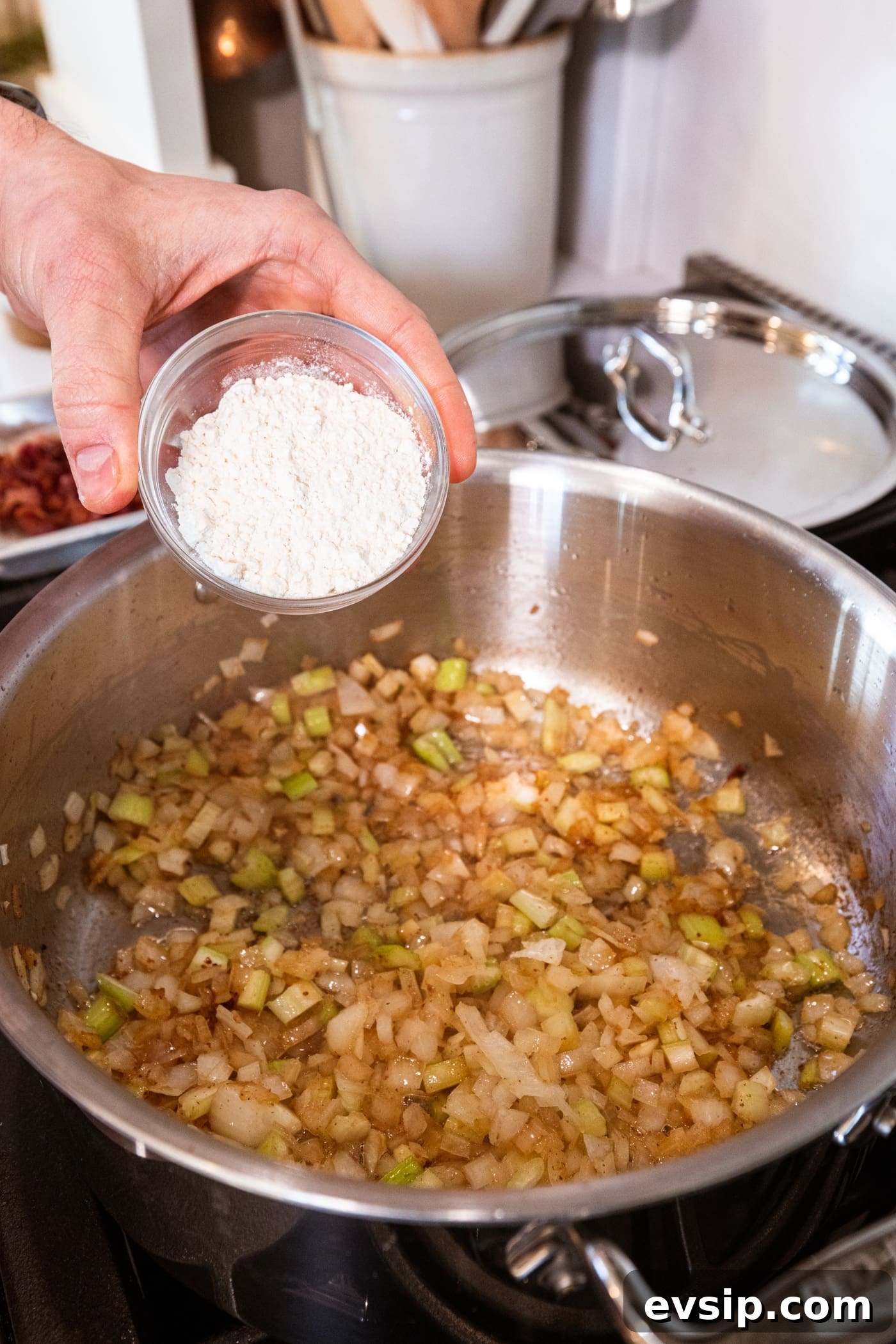 Hand about to pour flour into a large stockpot filled with cooked onions and celery.